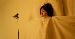 Woman lying wide awake in bed under warm lamp light, clutching the blanket, illustrating perimenopause insomnia